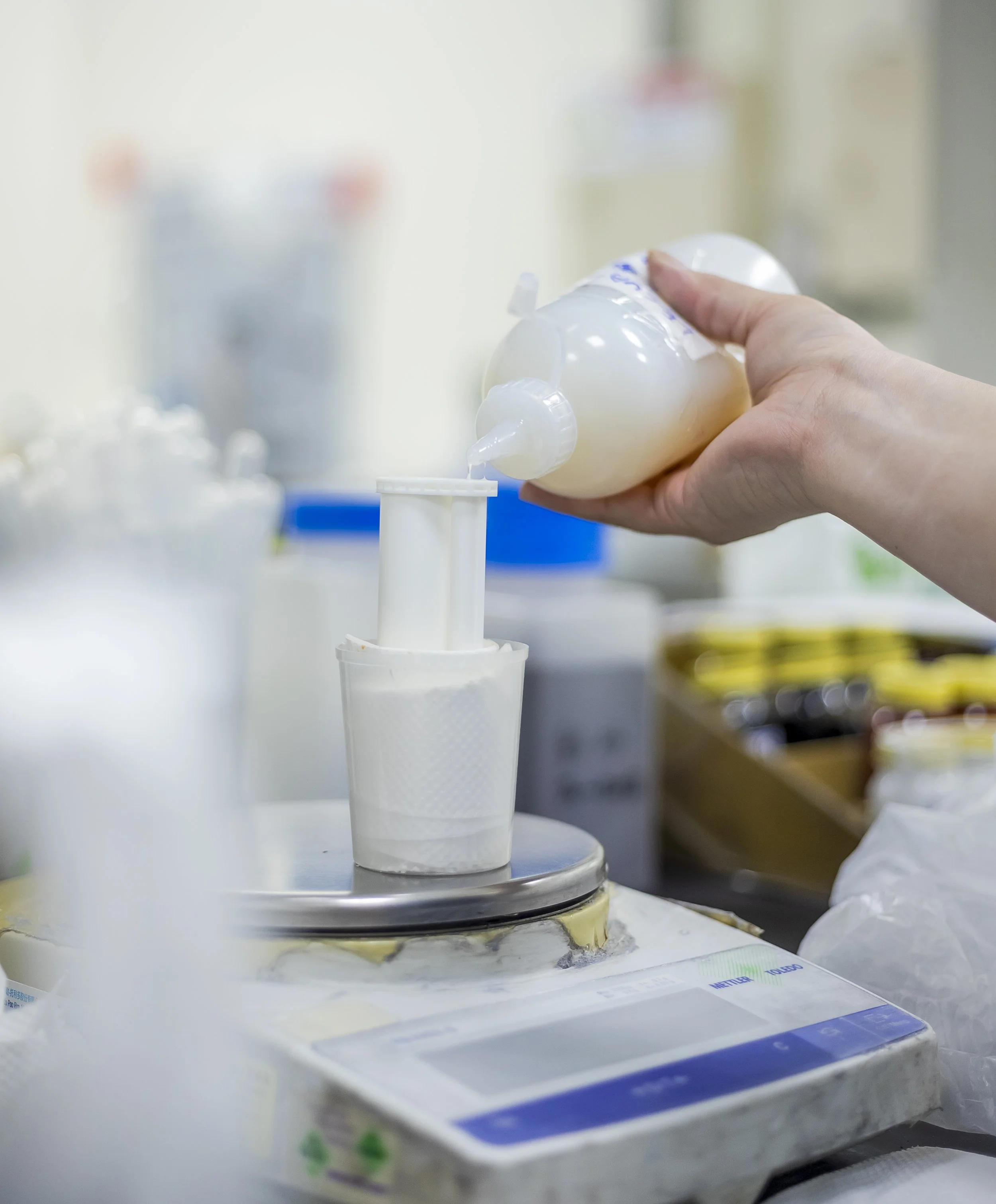 A hand pouring a creamy white substance into a cylindrical container on a scale in a laboratory or manufacturing setting.