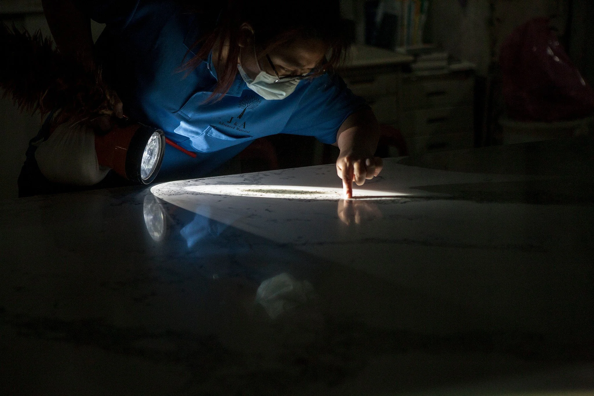 Person inspecting a large white surface with a flashlight in a dark room, wearing a face mask and a blue shirt.