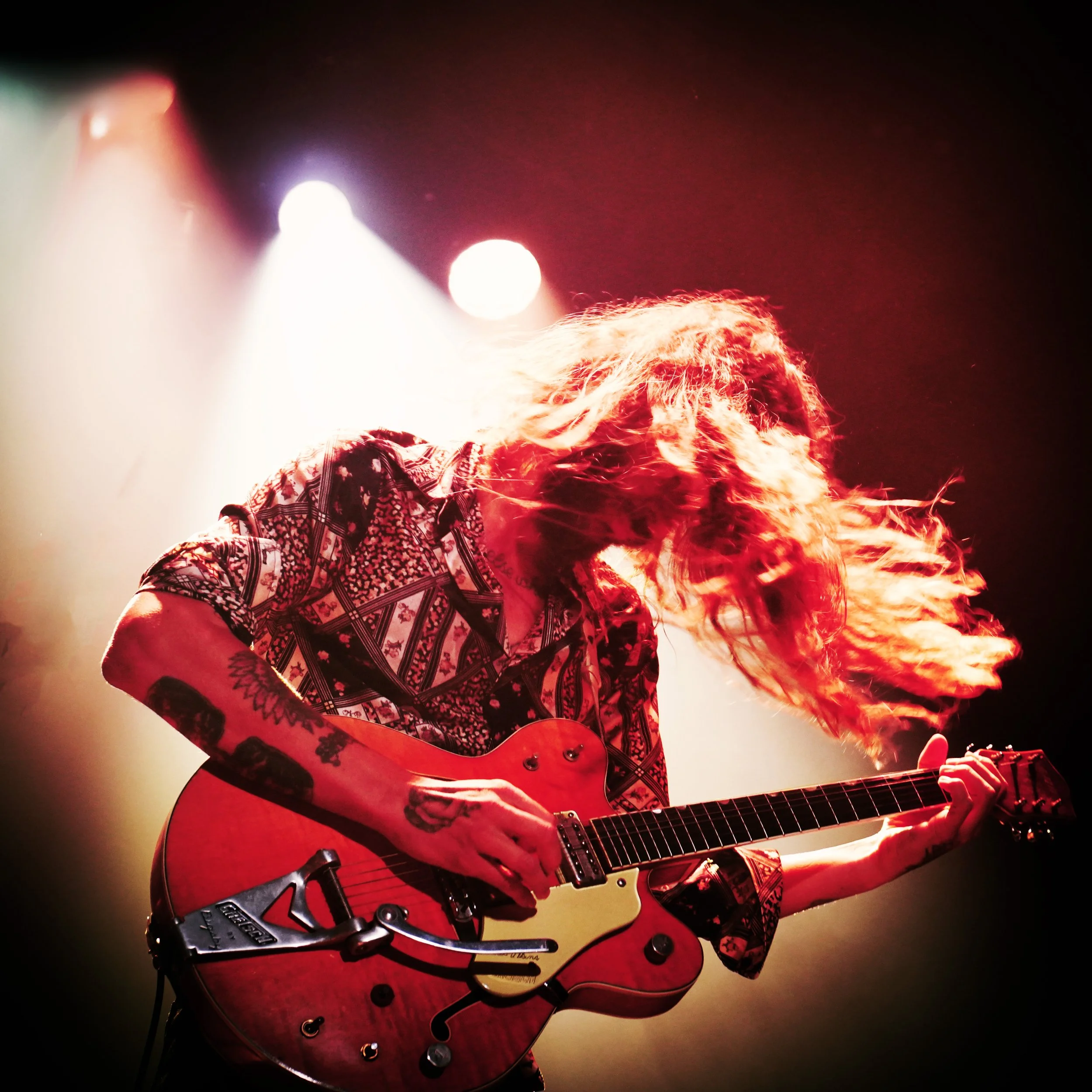 A person playing an electric guitar, with long hair covering their face, in a dark stage setting with red lighting.