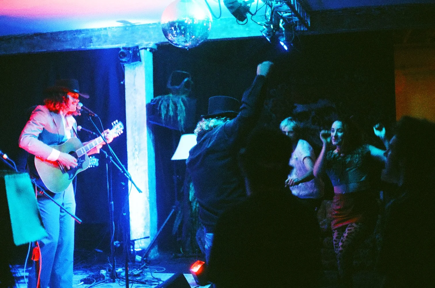A musician wearing a hat is playing an acoustic guitar and singing on stage, with a group of people dancing and enjoying the music under colorful lighting.