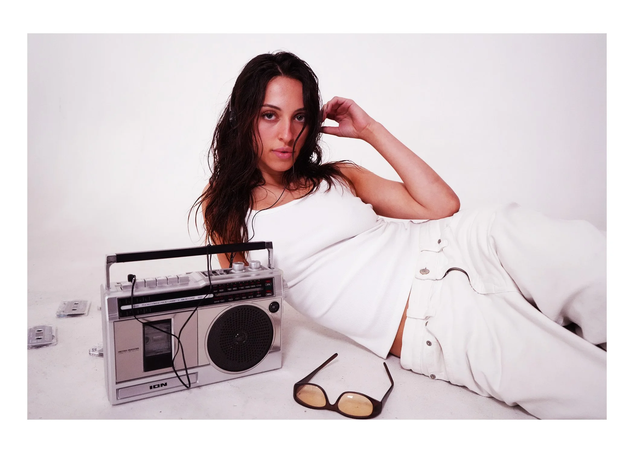 Woman with wet hair lying on white surface, wearing white shirt, with stereo radio, sunglasses, and electronic devices nearby, against plain white background.
