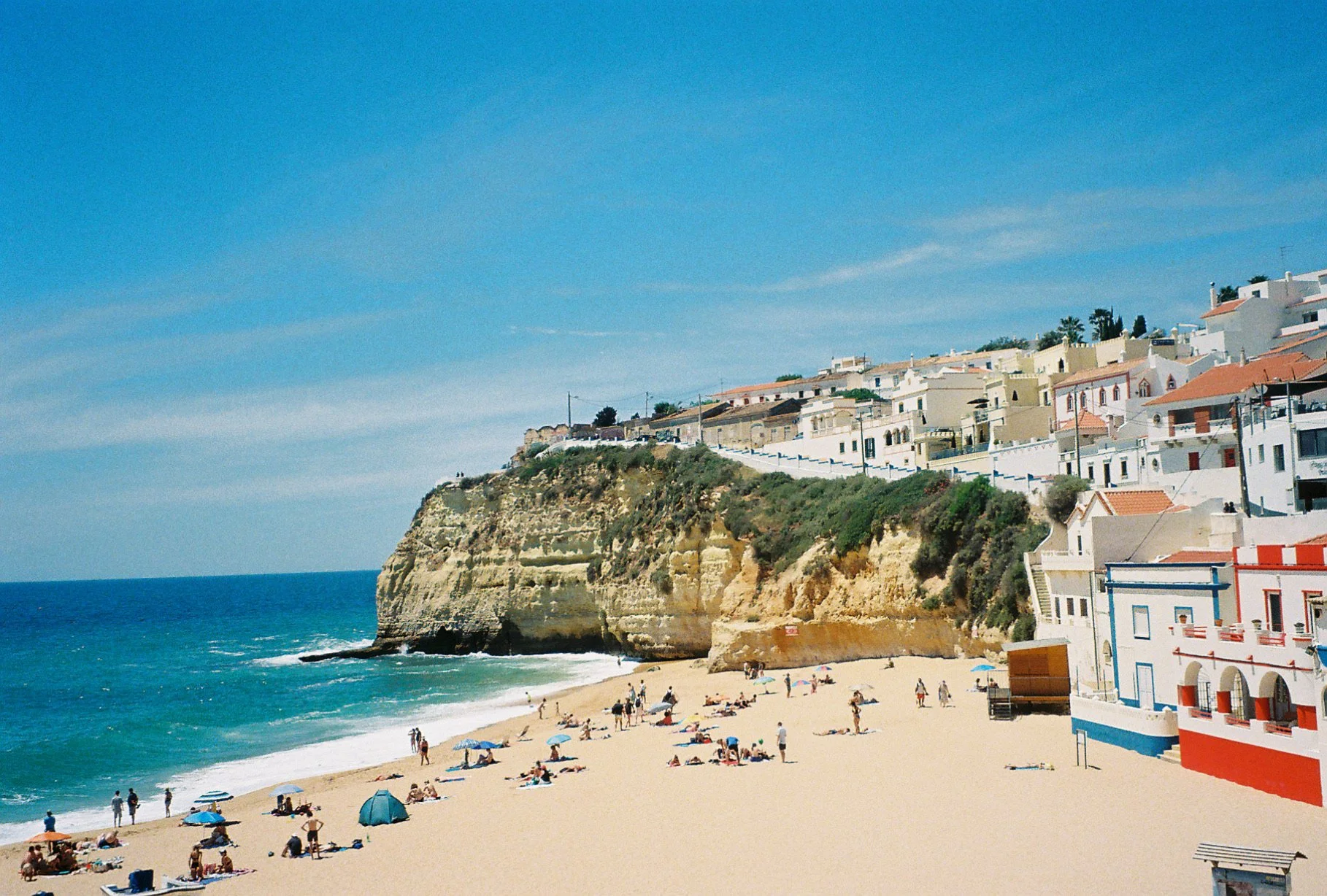Sunny beach with people relaxing under umbrellas, a cliff with white houses on top, and a clear blue sky.