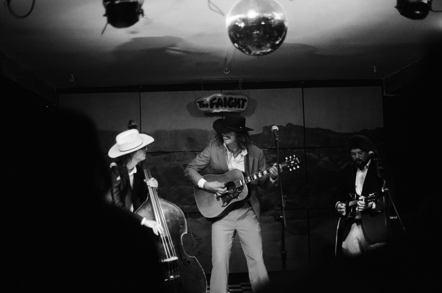 Three musicians performing on stage with guitars and a double bass, wearing cowboy hats, in a venue with a mountain landscape backdrop and a sign that reads 'The Faint'.
