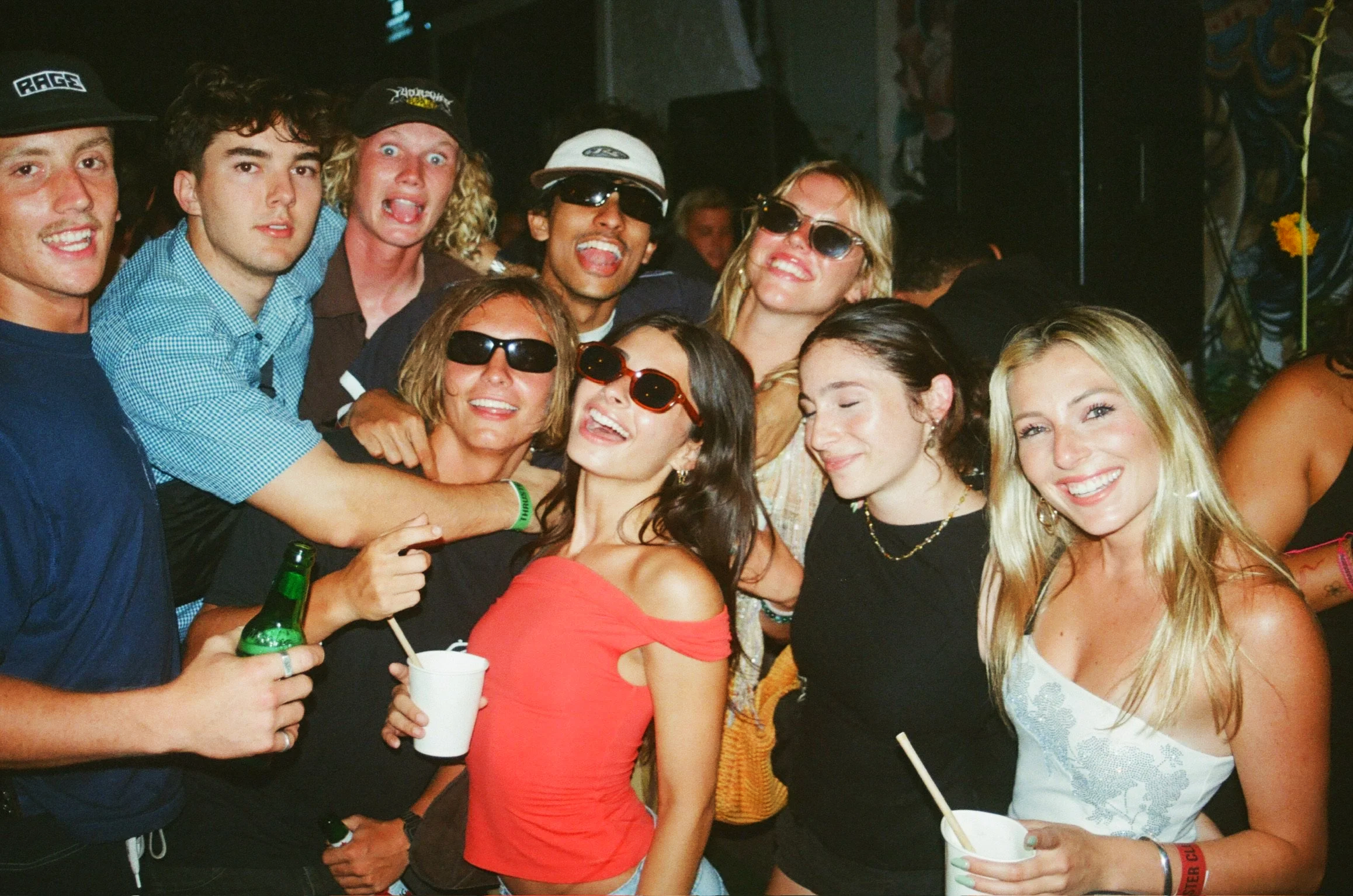 A group of friends at a party, smiling and posing for the camera, some wearing sunglasses, holding drinks, with a dimly lit background.