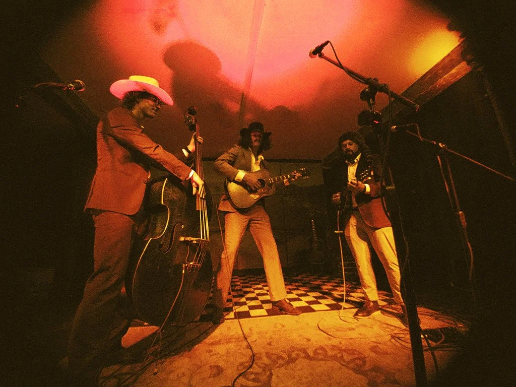 Three musicians performing on a small stage, playing guitars and a double bass, with dim orange lighting creating a cozy atmosphere.