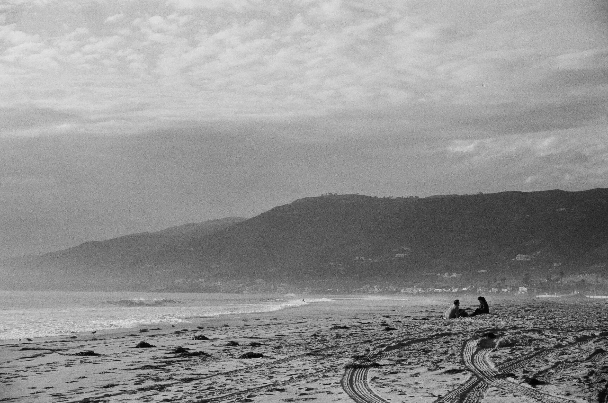 A black-and-white photo of a beach with two people sitting on the sand near the shoreline. Tire tracks are visible in the sand, and mountains with scattered trees are in the background under a cloudy sky.
