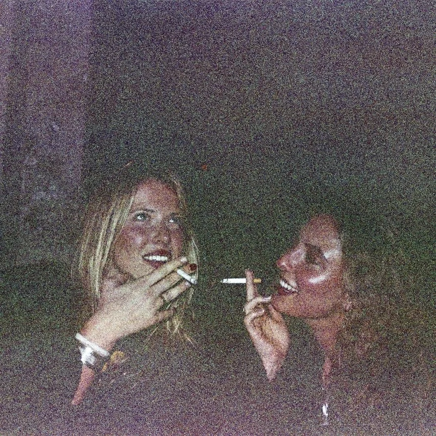 Two young women talking and smoking cigarettes at night, in a dark outdoor setting.