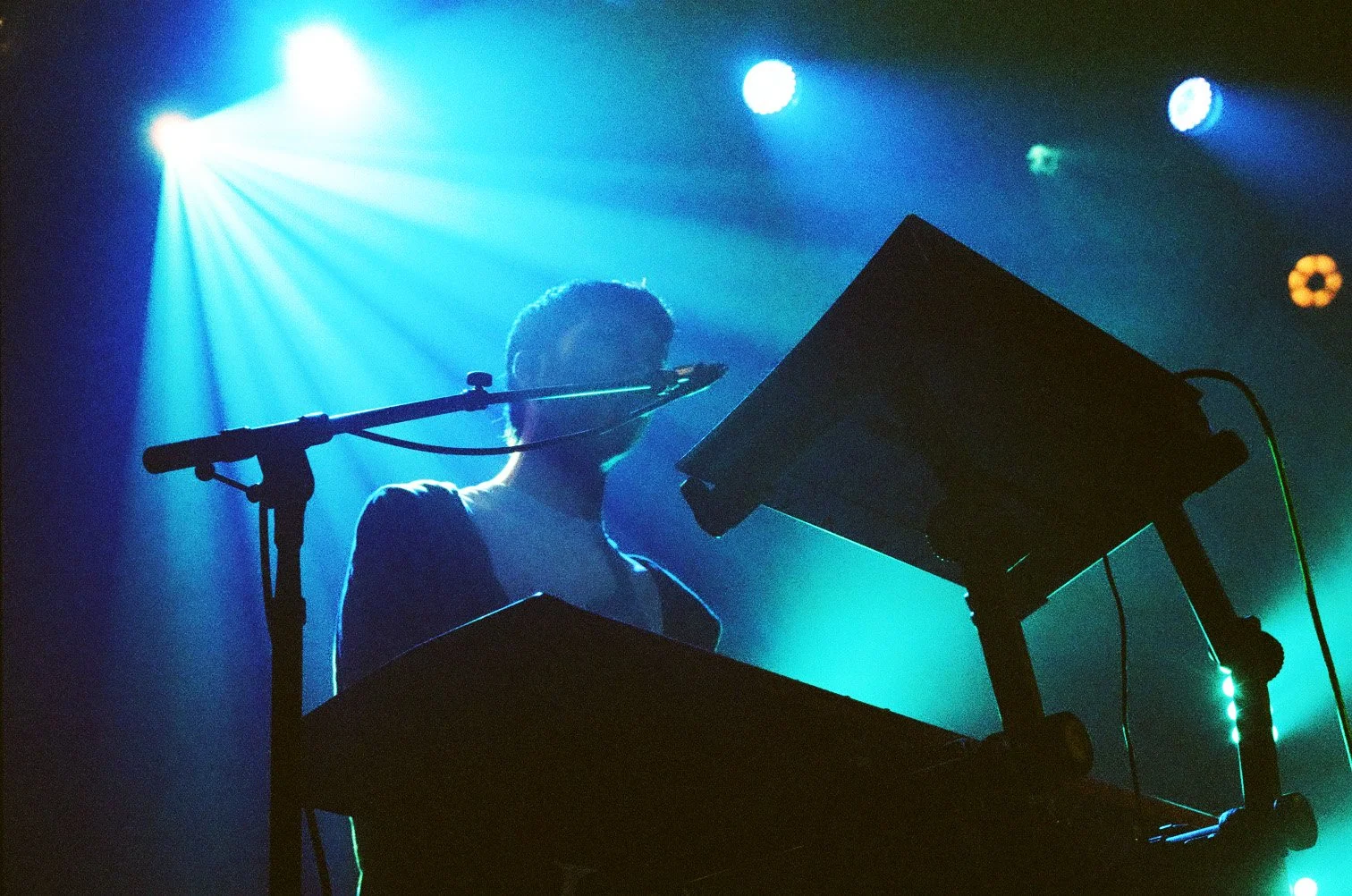 A silhouette of a musician standing behind electronic music equipment on a stage with blue and green lighting, with bright stage lights shining down from above.