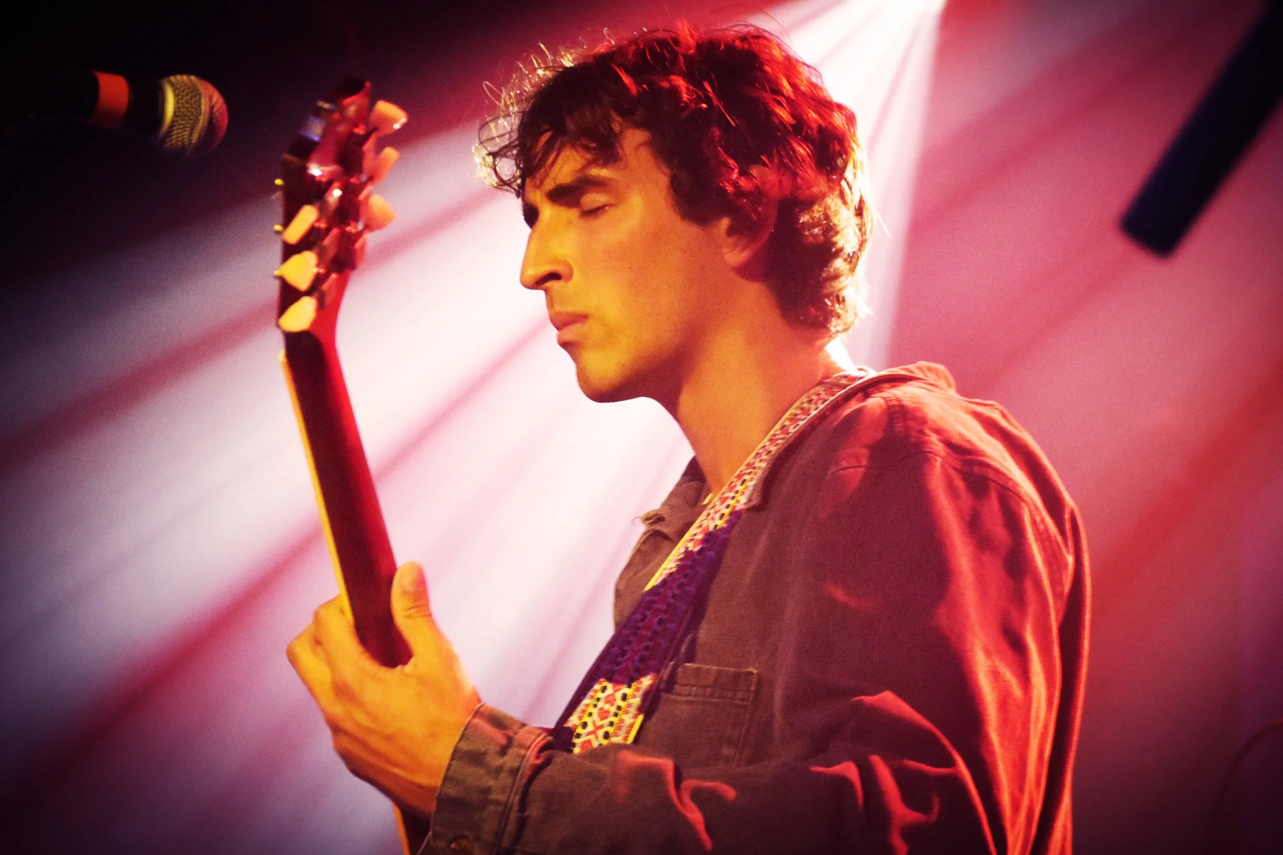 A young man with curly hair, eyes closed, plays an electric guitar in a dimly lit stage with pink and purple lighting.