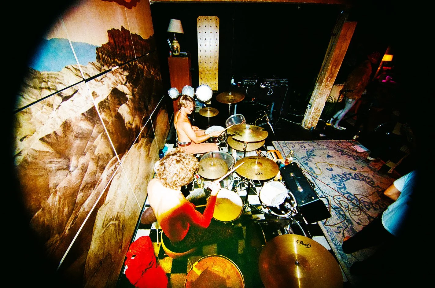 Two musicians performing on a small stage, one playing drums and the other singing, with a wall mural of mountains behind them and a carpeted floor with a patterned rug.