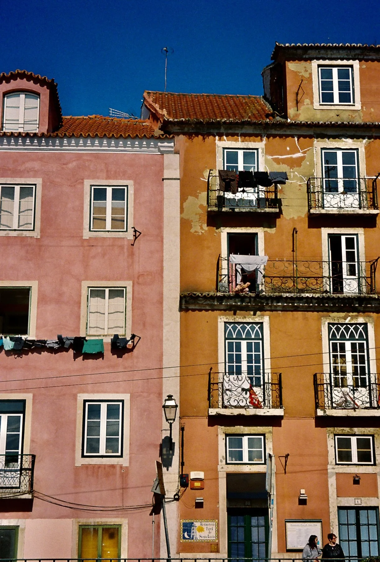 Colorful multi-story buildings with shuttered windows and balconies, laundry hanging outside, and a few people walking at the sidewalk in front, under a clear blue sky.