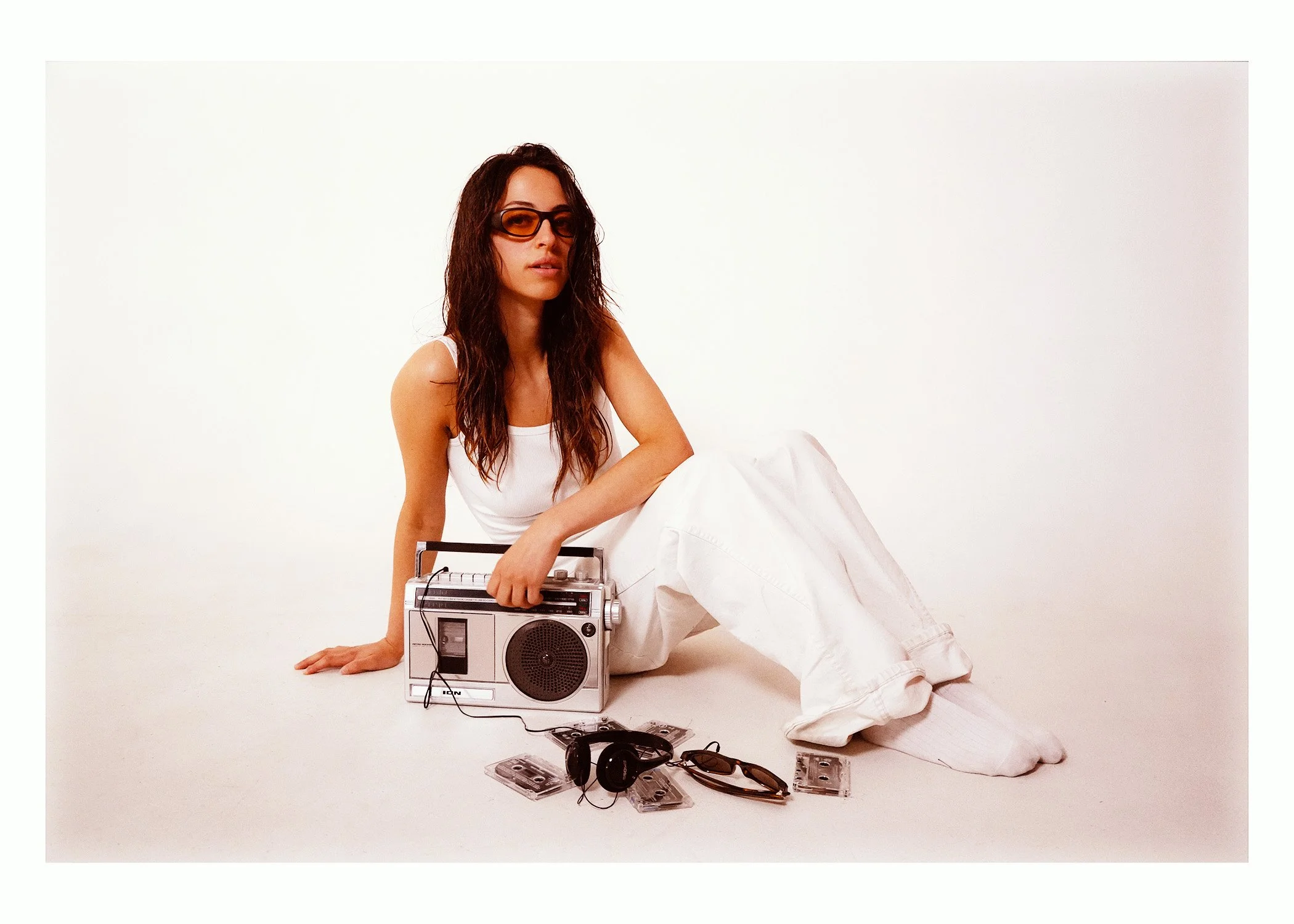 A woman with long brown hair, wearing sunglasses, a white tank top, and white pants, sits on the floor against a plain white background. She is holding a vintage boombox, with several cassette tapes, sunglasses, and headphones placed on the ground ar