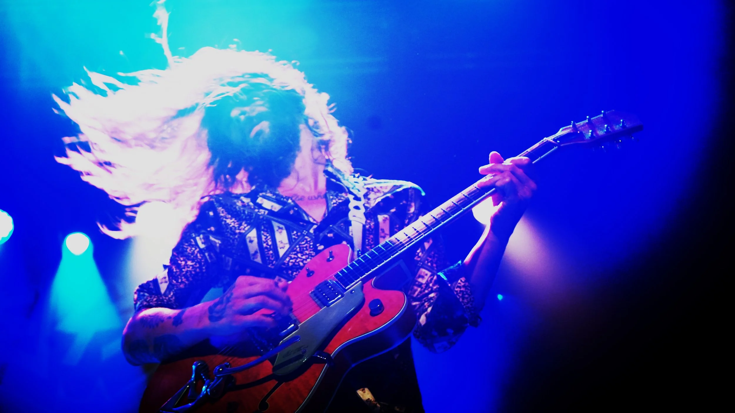 A female guitarist performing on stage under colorful lights with her hair flying, wearing a patterned jacket and playing an electric guitar.
