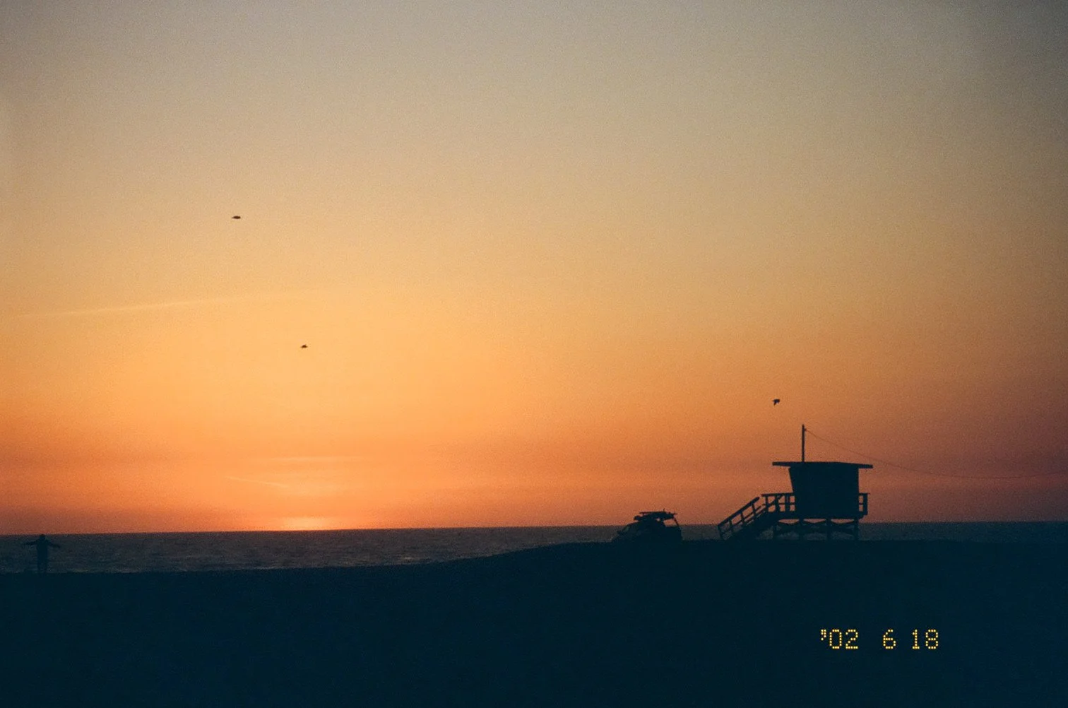 A sunset over the ocean with a lifeguard tower and a boat on the beach, birds flying in the sky.