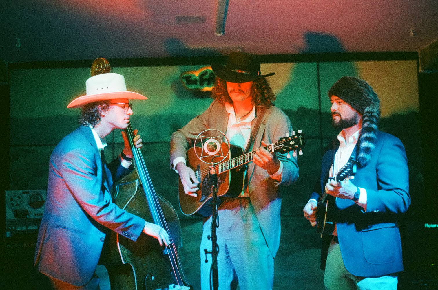 Three musicians performing on stage with colorful lighting, wearing distinctive hats and suits, holding guitars, with a green landscape backdrop.