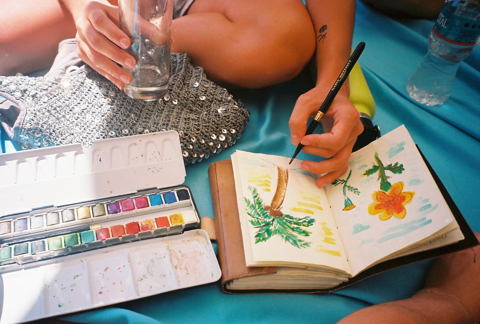A person sitting on a teal surface, painting a colorful landscape in a sketchbook. The person is holding a black pen and is surrounded by watercolor paints, a water glass, a water bottle, and a sequined bag.