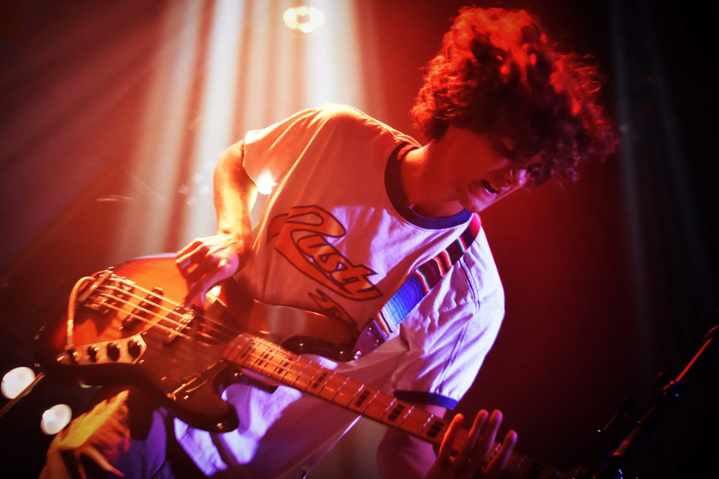 A musician with curly hair playing an electric guitar on stage, illuminated by red and orange stage lights.