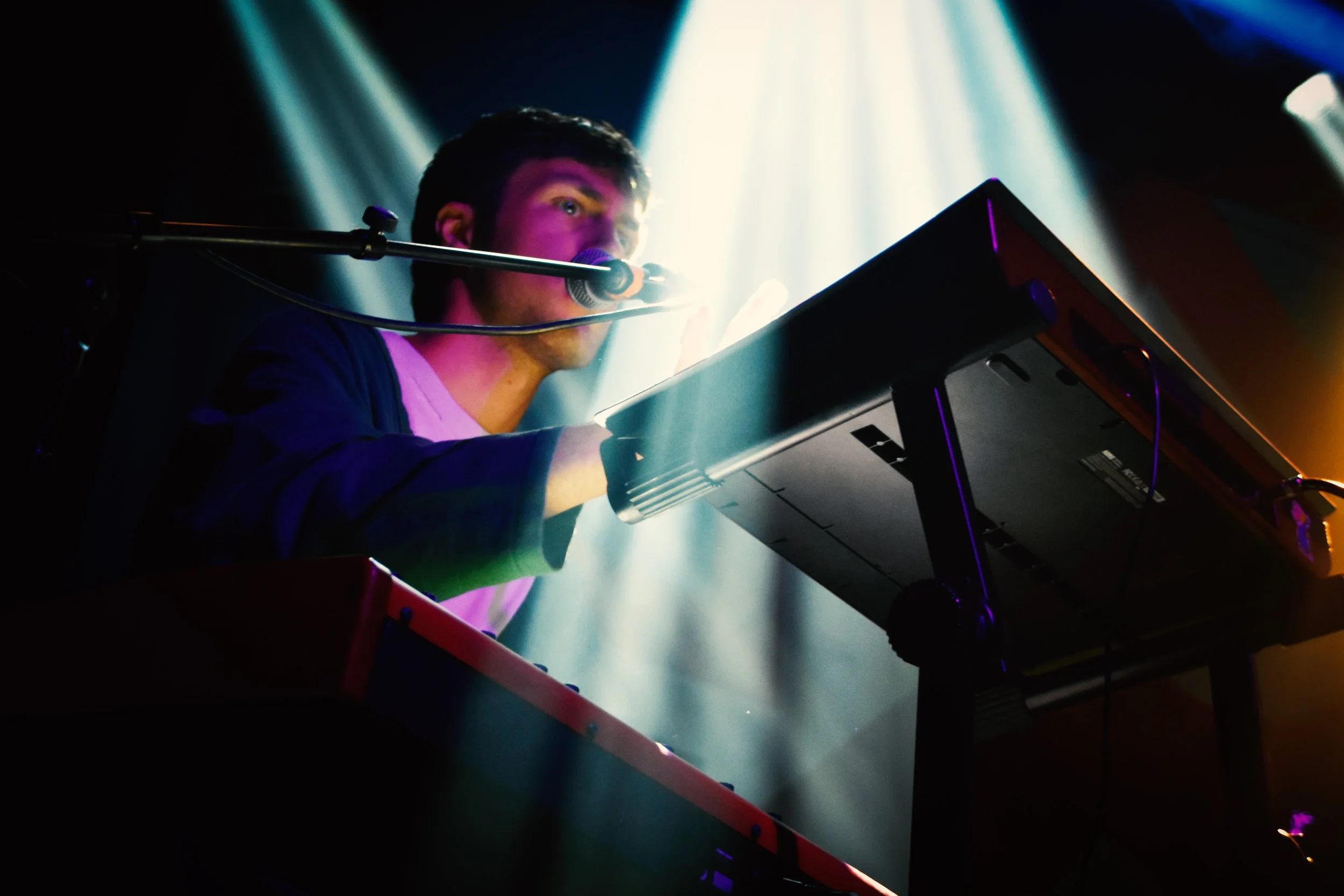 A man performing on stage with a keyboard in front of him, illuminated by colorful stage lights.