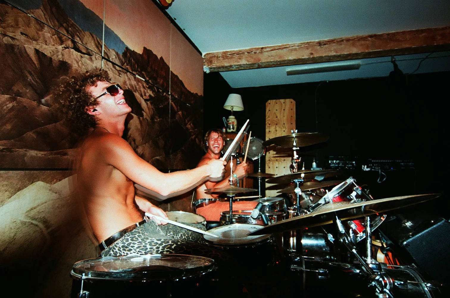 Two shirtless men playing drums and singing, one wearing sunglasses and leopard print pants, in a room with a mountain mural on the wall.