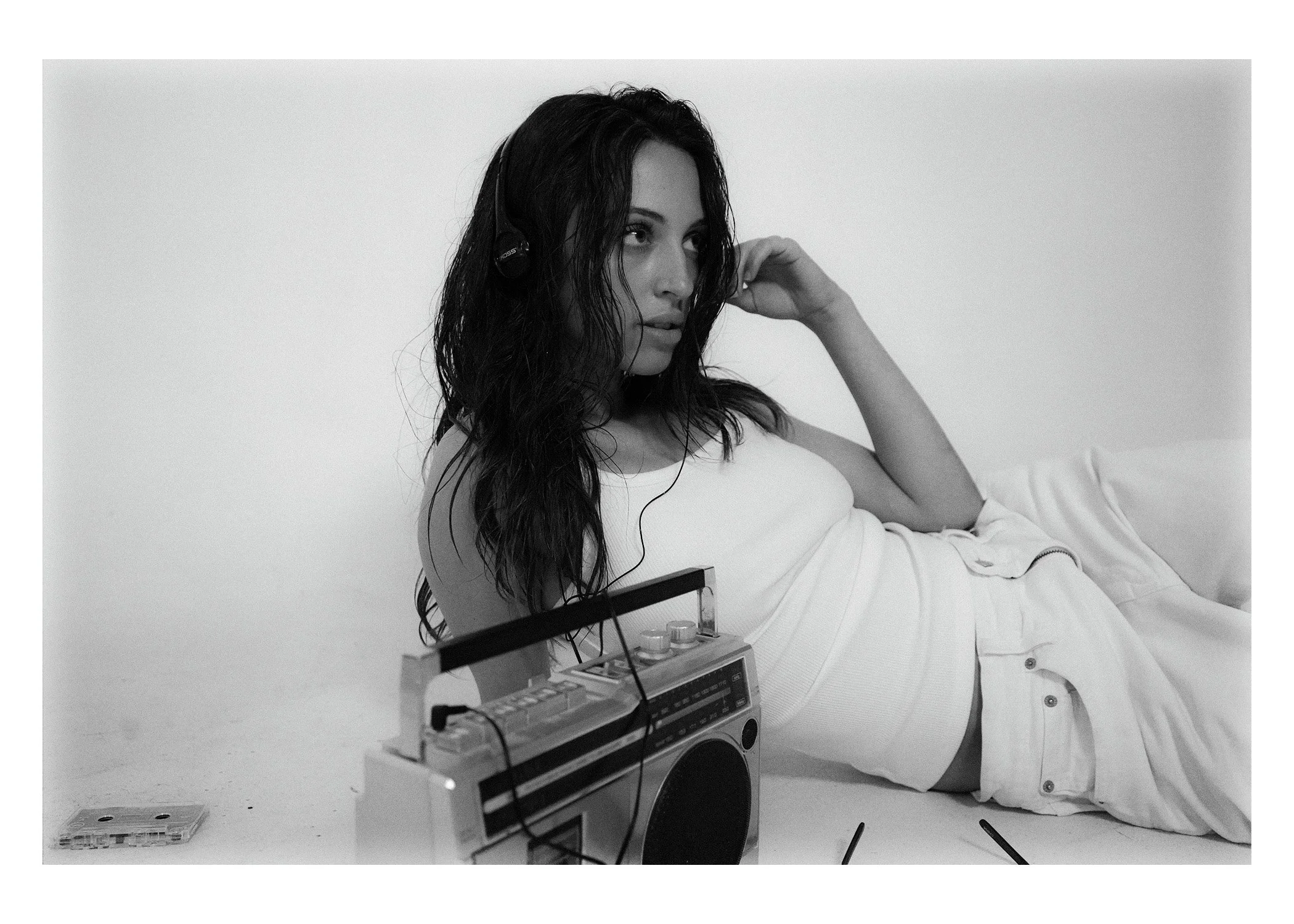 A woman with wet hair wearing headphones, lying on a bed with an old-fashioned stereo system and a cassette tape player nearby, in a minimalist room.