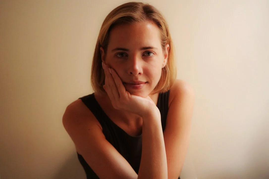 A woman with short blonde hair and fair skin resting her face on her hand, wearing a sleeveless black top, against a plain beige wall.