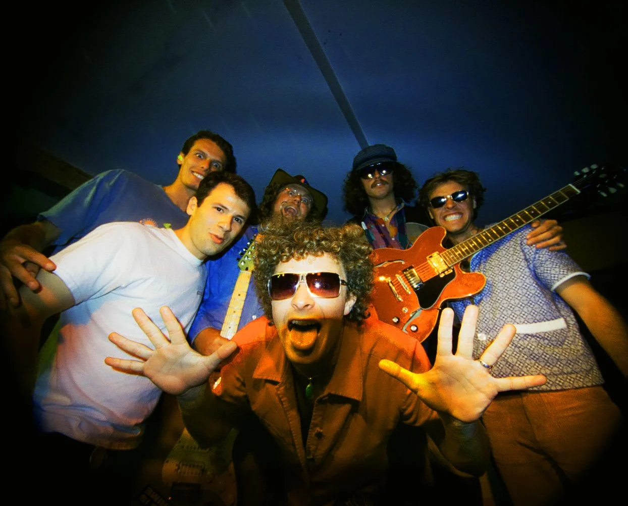 Group of six men at a party or concert, some holding guitars, making fun expressions, one wearing sunglasses, in a dimly lit indoor setting.