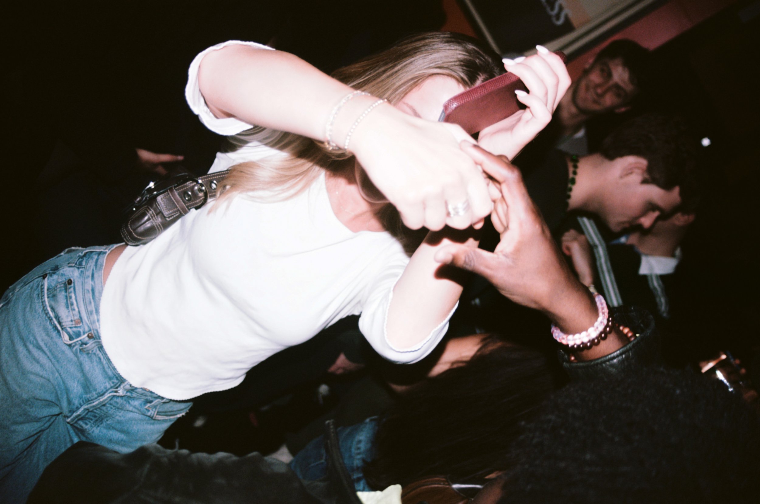 People dancing closely at a party with a woman holding a phone, wearing casual clothes and jewelry, and several others visible in the background.