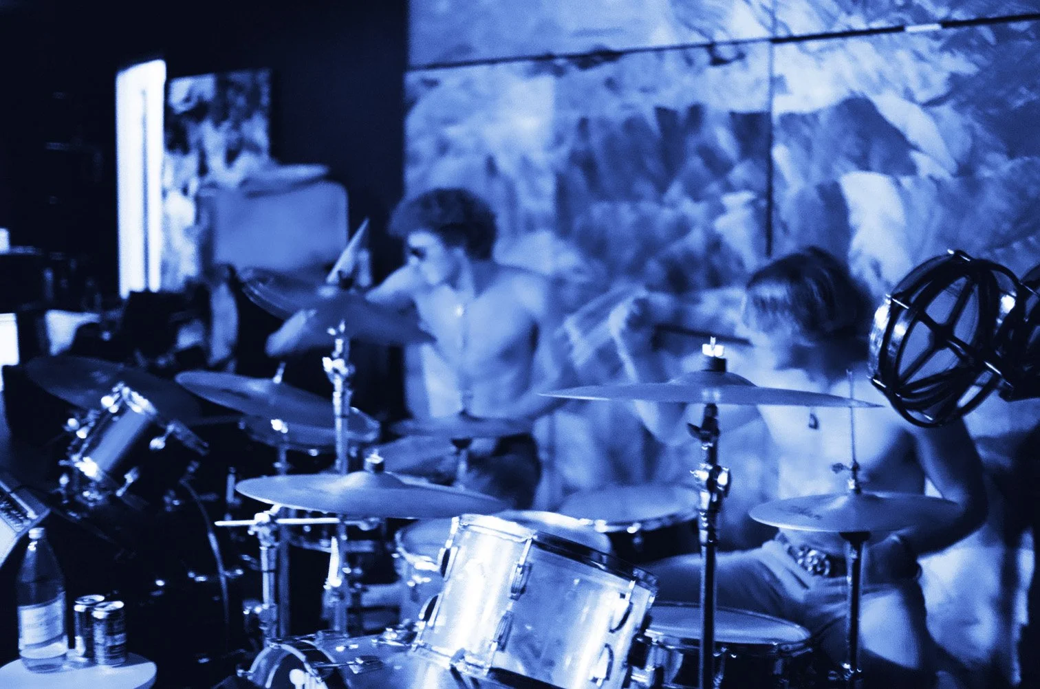 Two shirtless musicians, one playing drums, performing in a dimly lit venue with a textured wall background.