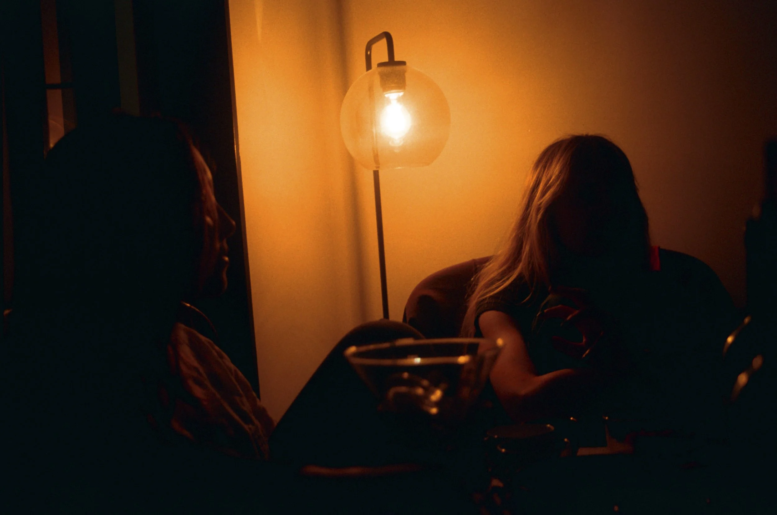 People sitting around a table in dim lighting, illuminated by a standing lamp with a glass shade.