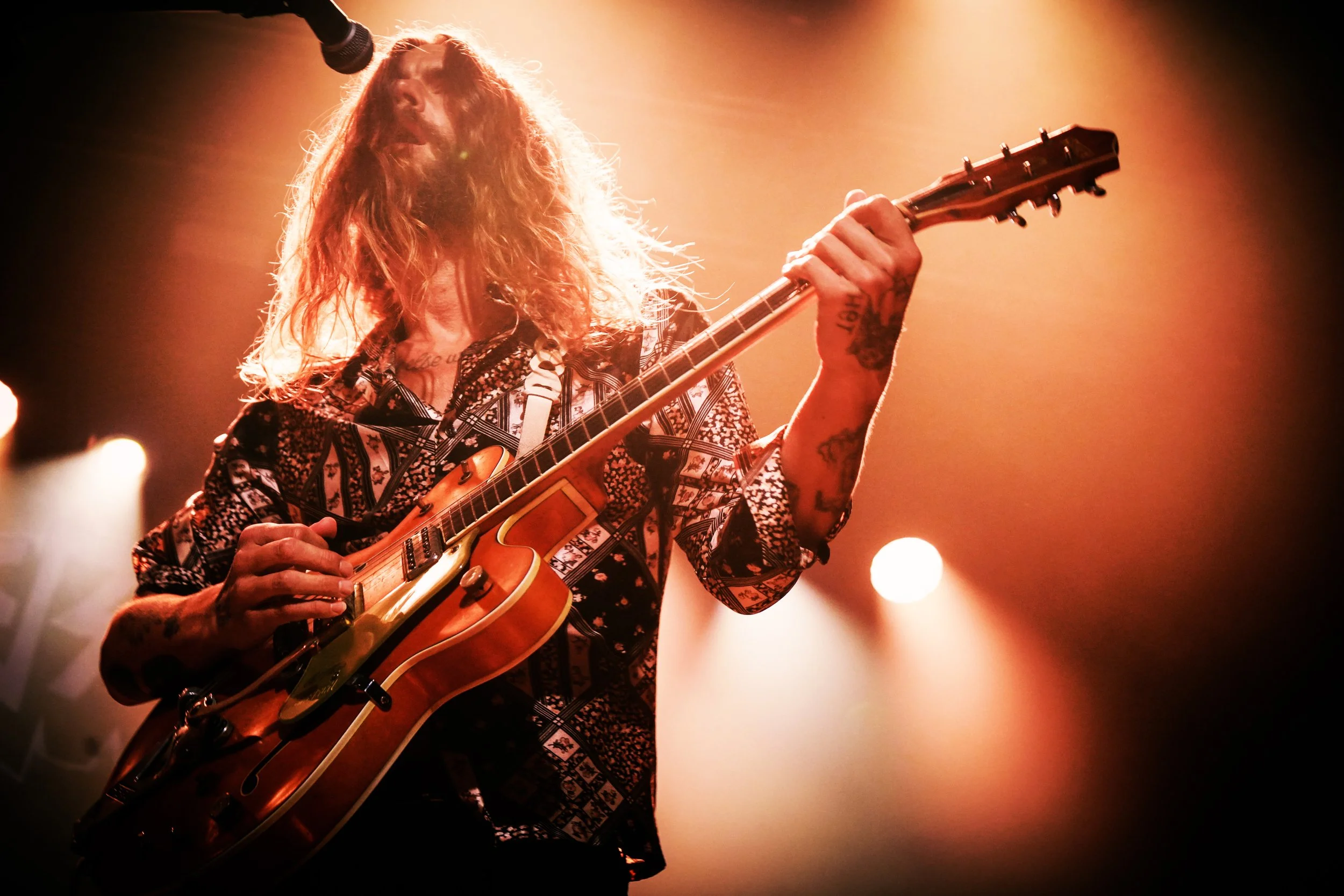 A musician with long, wavy hair playing an electric guitar on stage under warm lighting.