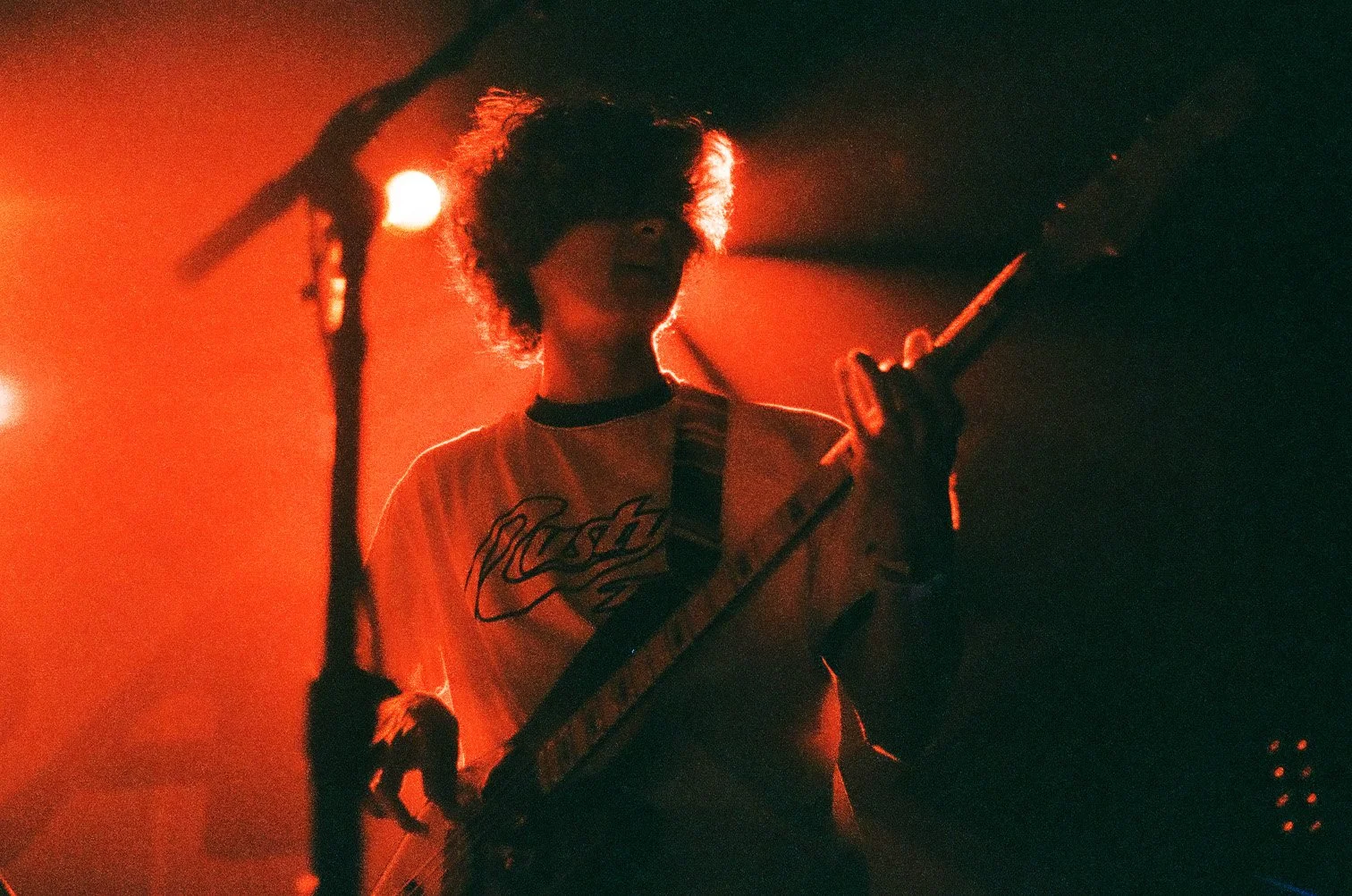A person with curly hair playing an electric guitar on stage, illuminated by red stage lights, with a microphone stand visible in the foreground.