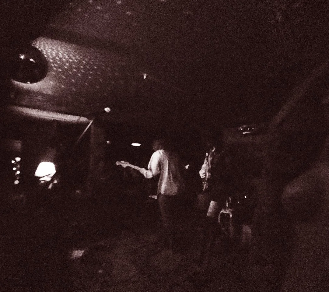 Two people playing guitars in a dimly lit room with a lamp and a television.