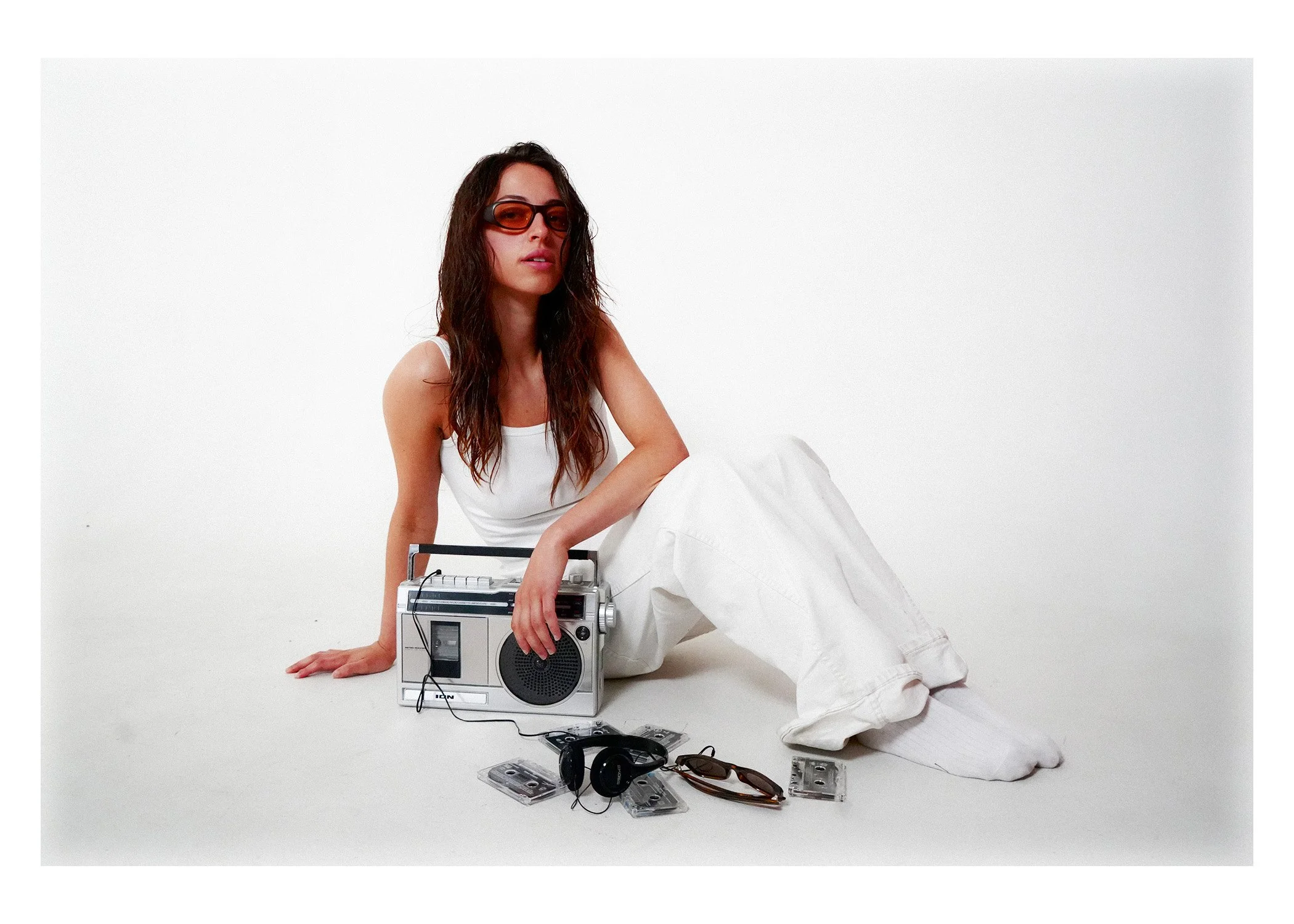 Woman in white clothing with long brown hair and sunglasses sitting on the floor surrounded by vintage audio equipment, cassette tapes, and accessories in a minimalist white setting.