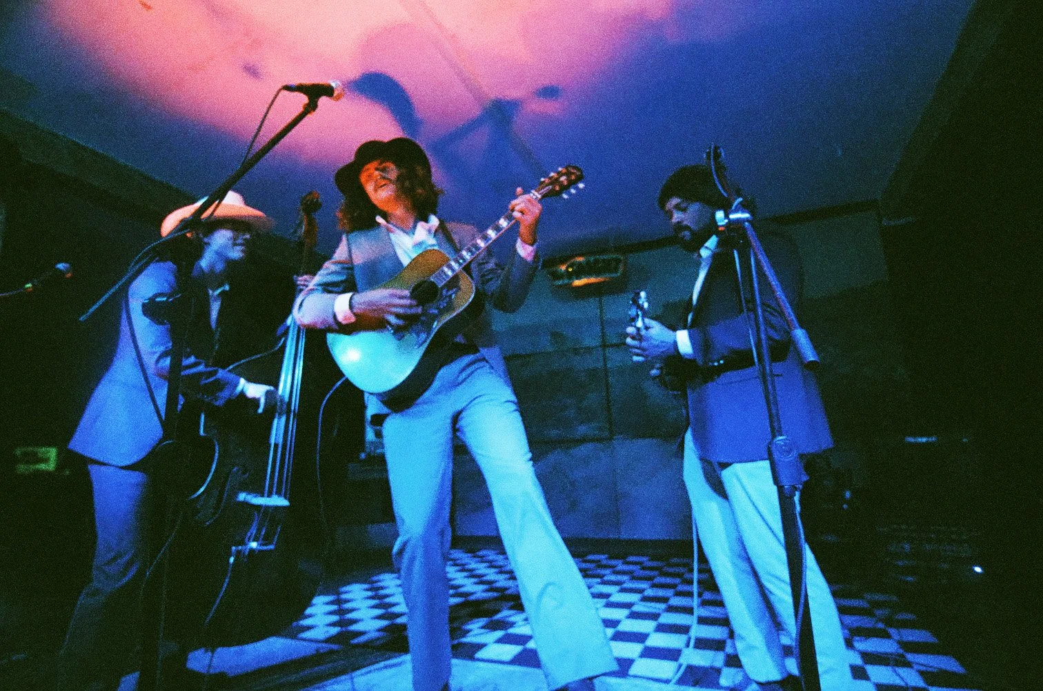 Musicians performing on a stage with one woman playing an acoustic guitar and singing, while the others hold microphones or musical equipment, in a dimly lit room with checkered flooring and colorful lighting.