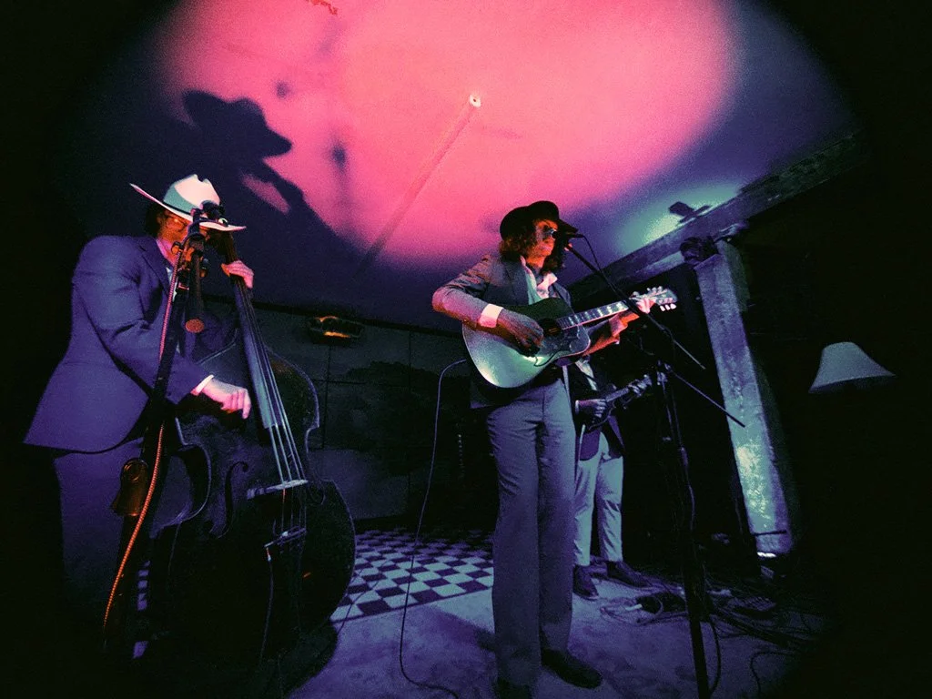 Musicians performing on a small stage, playing guitars and an upright bass, under dim purple and blue lighting.