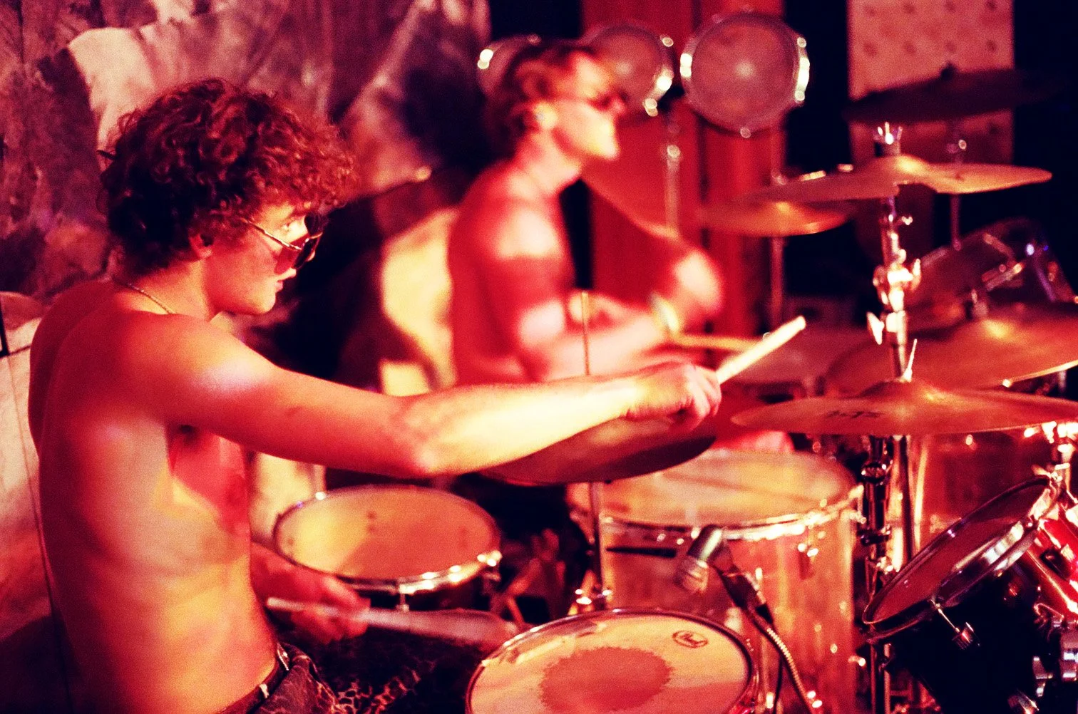 Two shirtless musicians playing drums in a dimly lit room with red lighting.