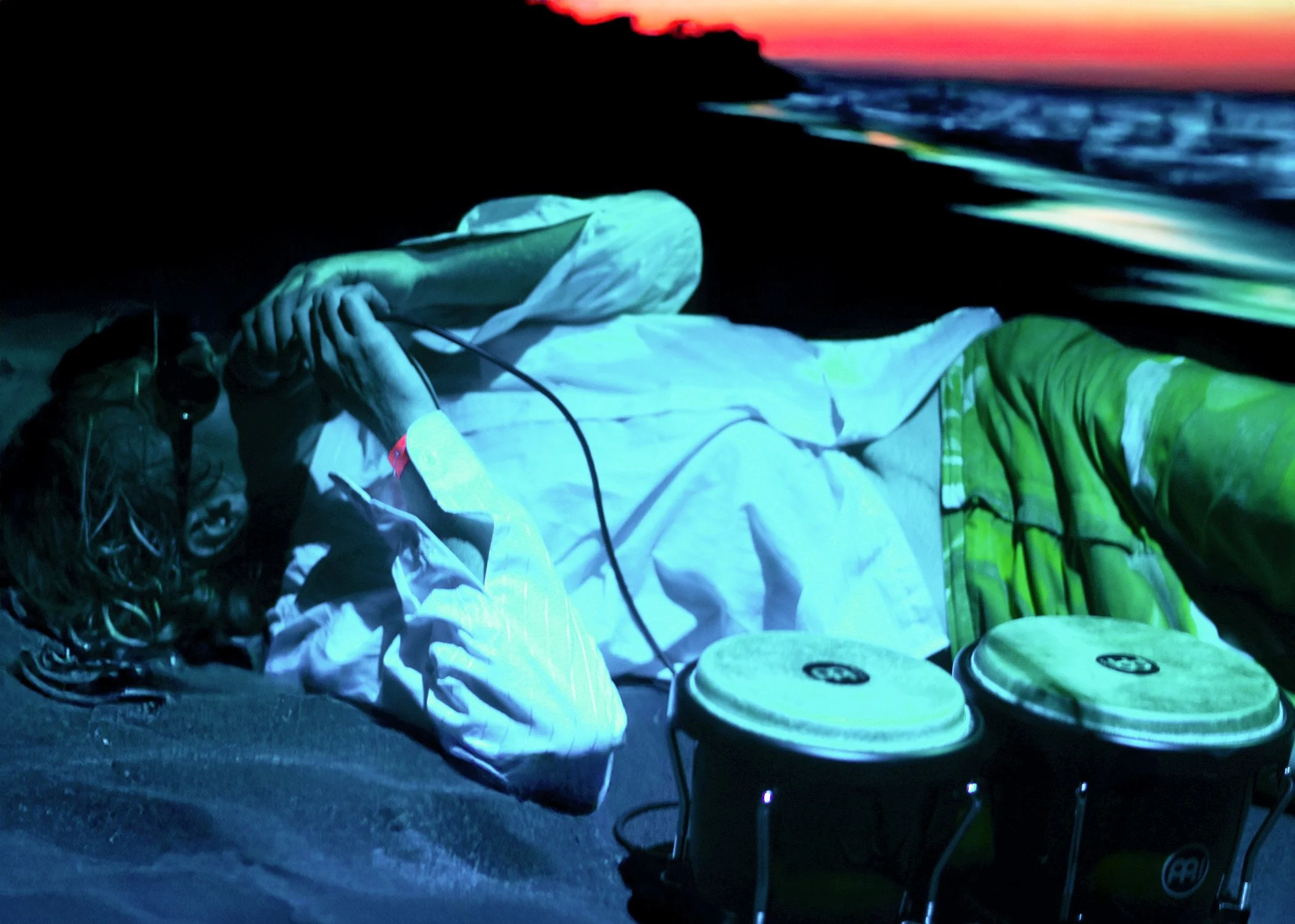 A person lying on the ground at sunset, with musical equipment including drum pads and cables nearby.