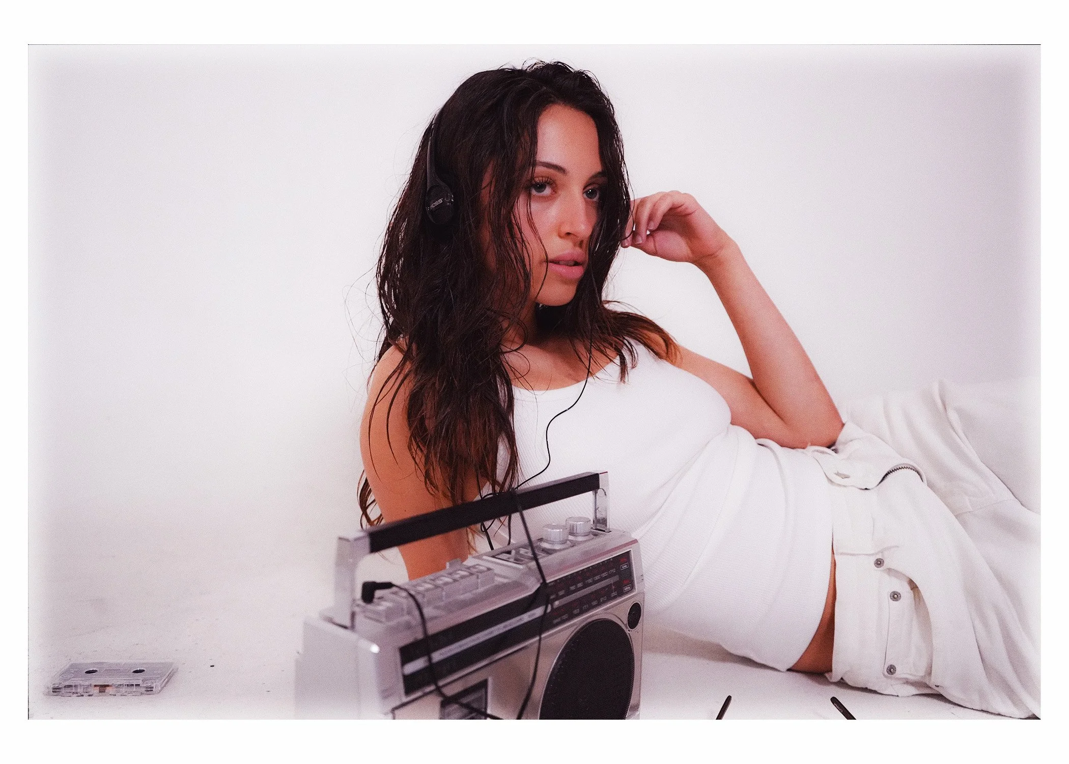 Young woman with curly dark hair listening to music through black headphones, holding her hand near her face, with a vintage boombox on a white surface in front of her and a small cassette tape to the side, all set against a white background.
