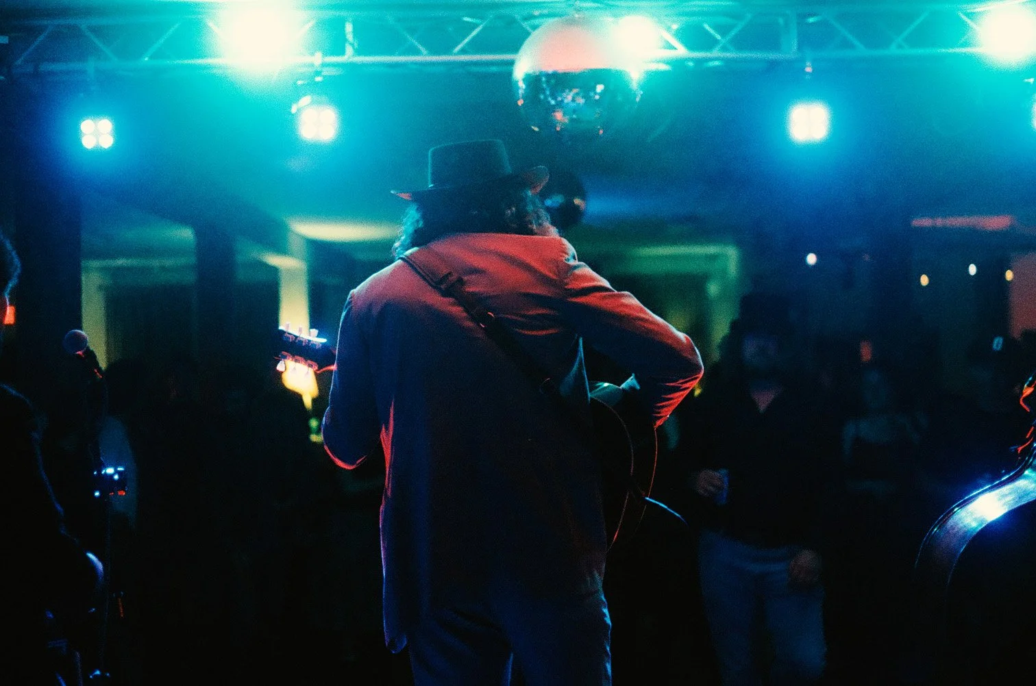 Performer playing guitar on stage with blue and green lighting, wearing a hat and reflective glasses, in a dark room filled with audience.