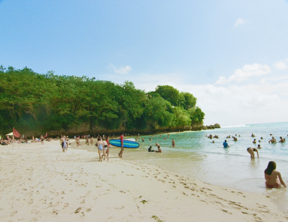 People swimming and relaxing on a sandy beach with green trees and rocky cliffs in the background under a blue sky