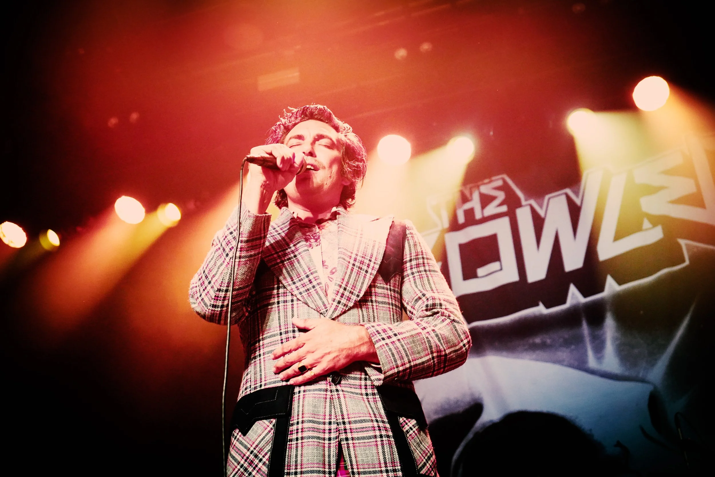A man with long hair and a checkered blazer singing into a microphone on stage with bright lights behind him.