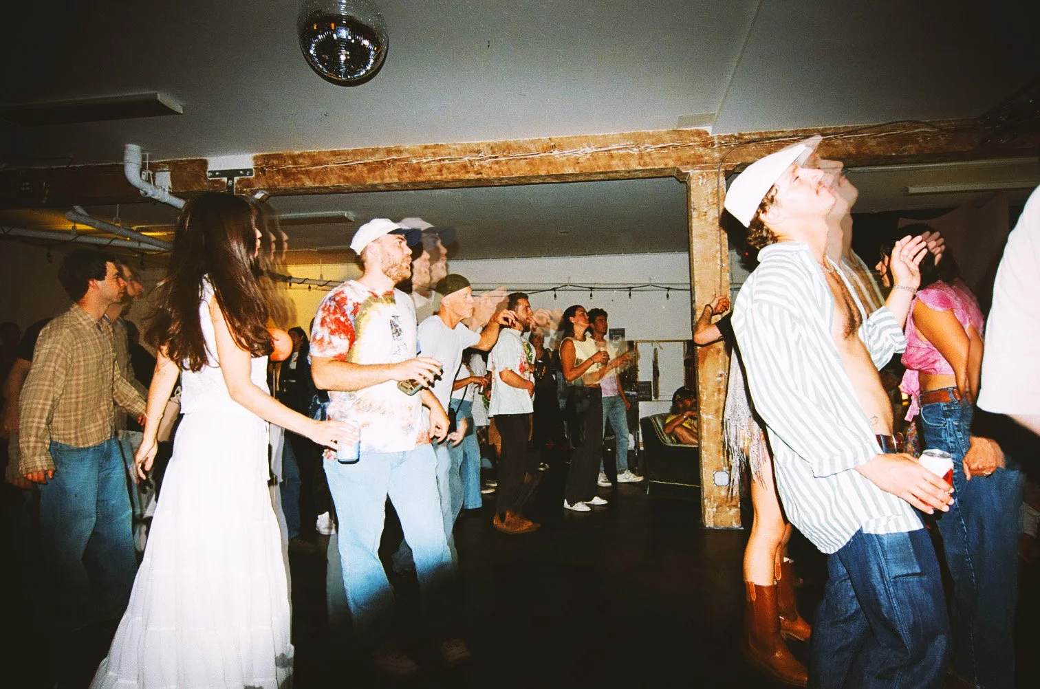 People attending a dance party or concert, dancing and holding drinks, in a dimly lit indoor venue with string lights and wooden beams.