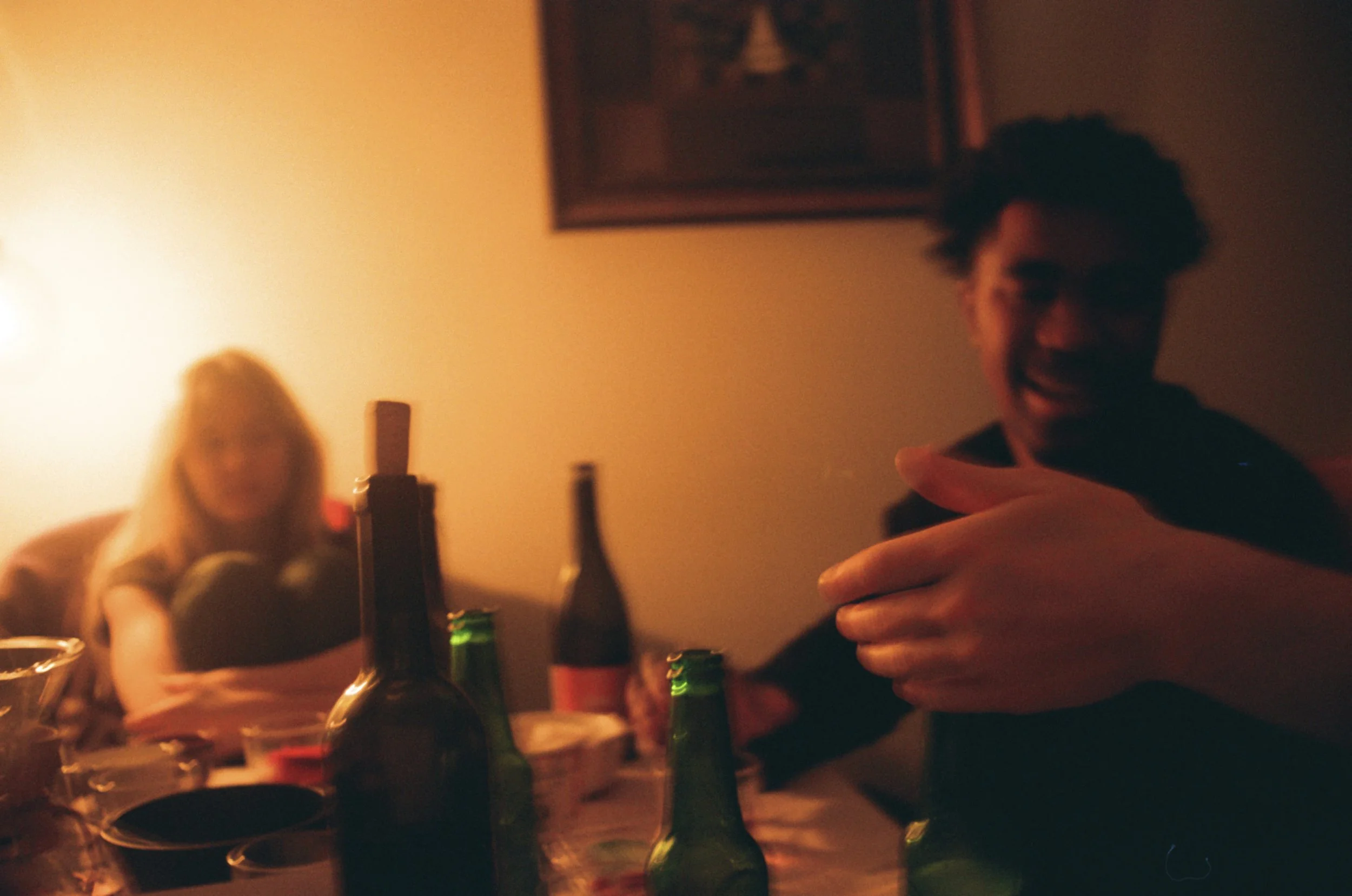 A person with curly hair taking a selfie in a dimly lit room, with several bottles on a table in front of them. A woman with long blonde hair is in the background sitting at the table, slightly out of focus.