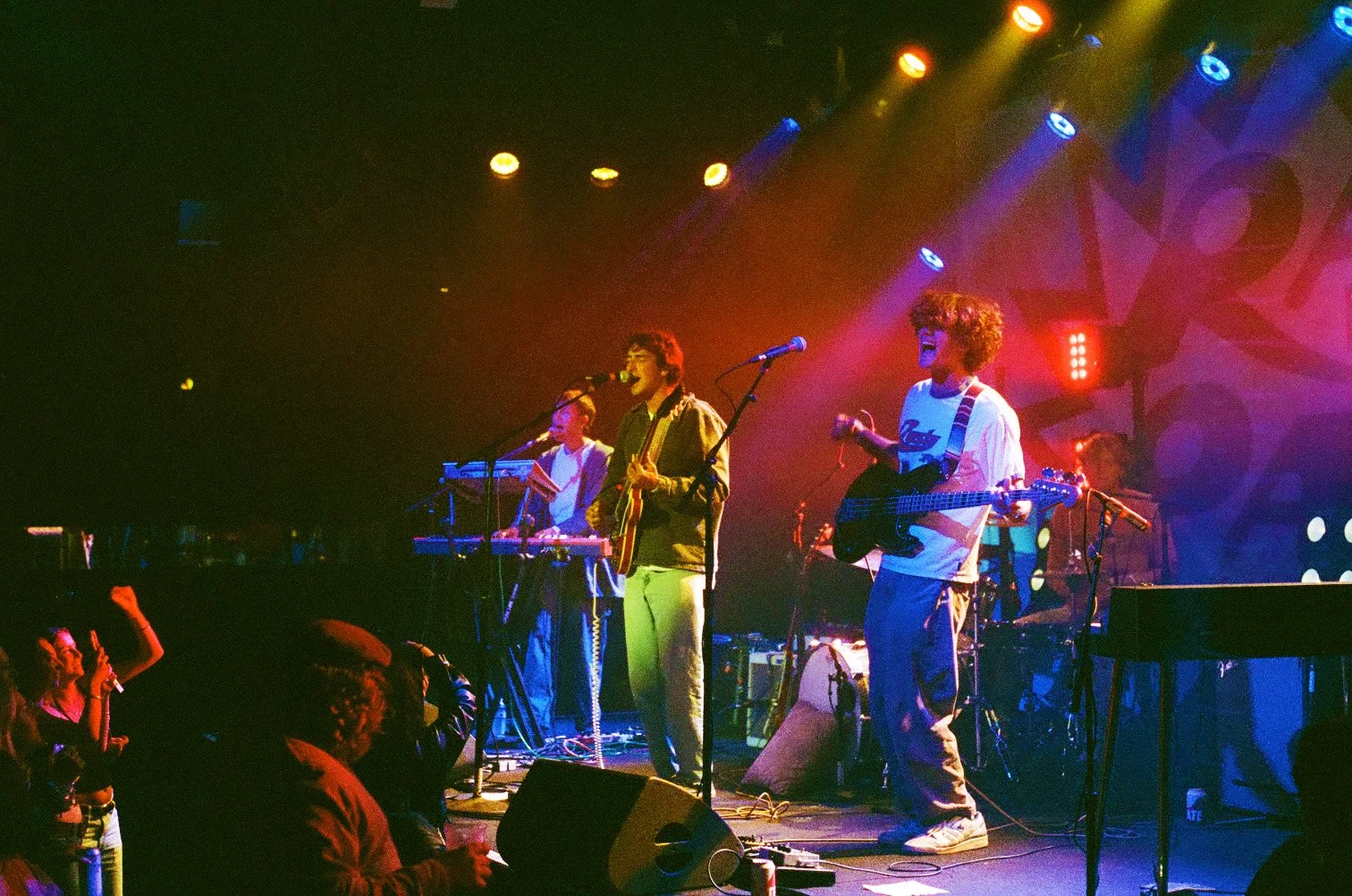 Band performing on stage with colorful lights, two members playing guitar and singing, audience in front enjoying the show.