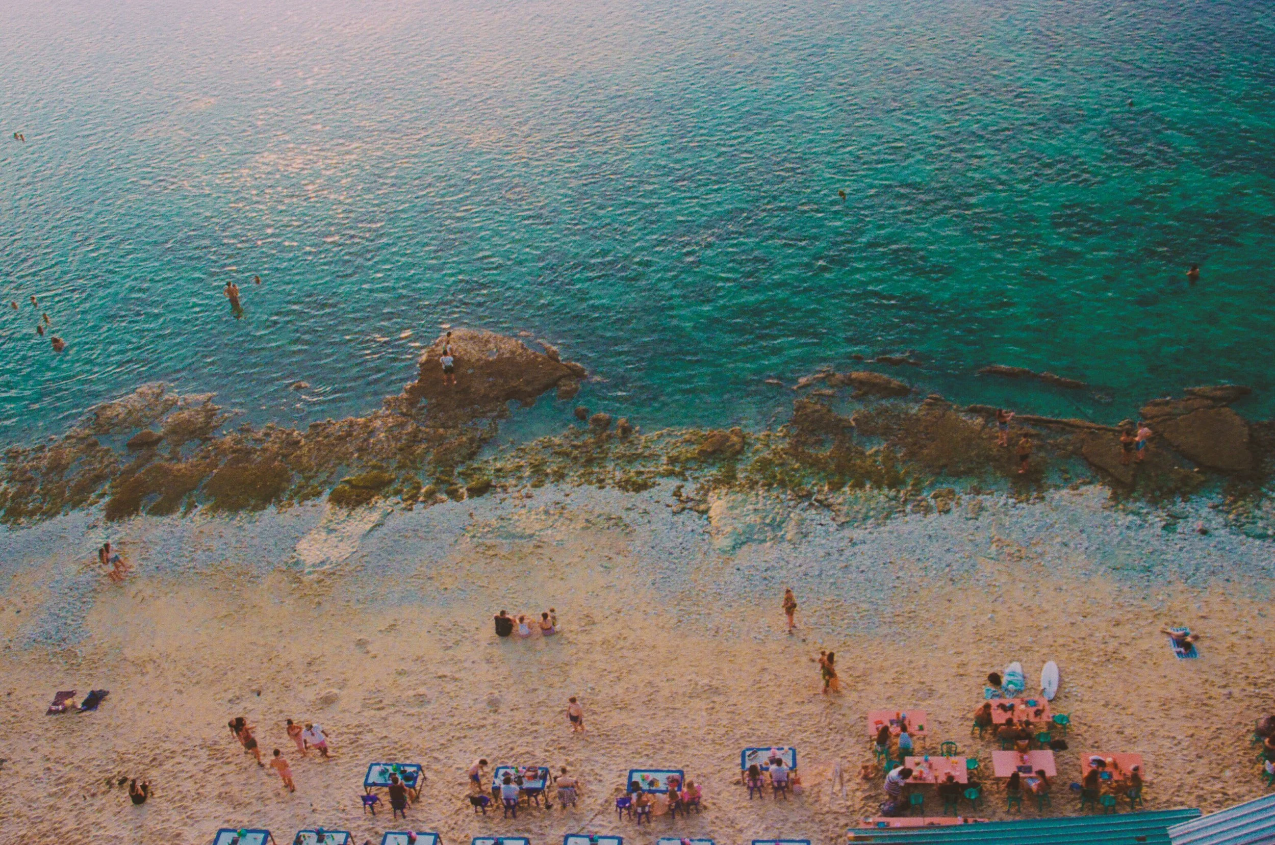 Beach with people sitting at tables on sand, some in water, and rocks along shoreline, with the ocean in the background.
