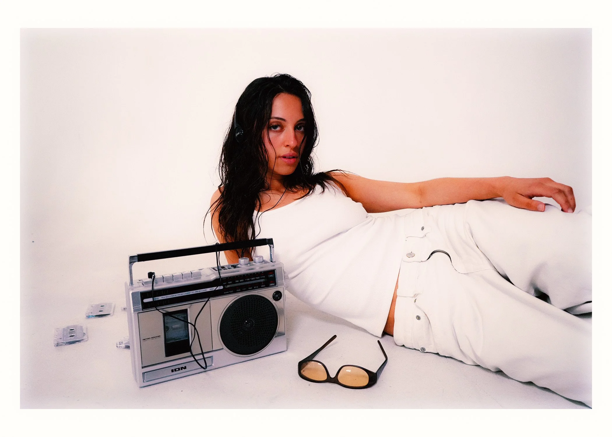 Woman lying on a white surface, wearing white clothing, with a vintage portable radio, sunglasses, and small electronic devices around her, against a plain white background.