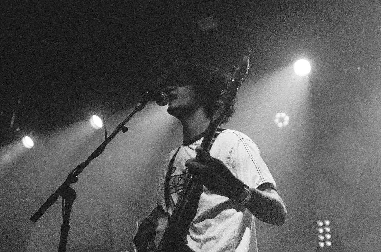 A black and white photo of a musician singing into a microphone on stage while playing an electric guitar, with stage lights shining in the background.