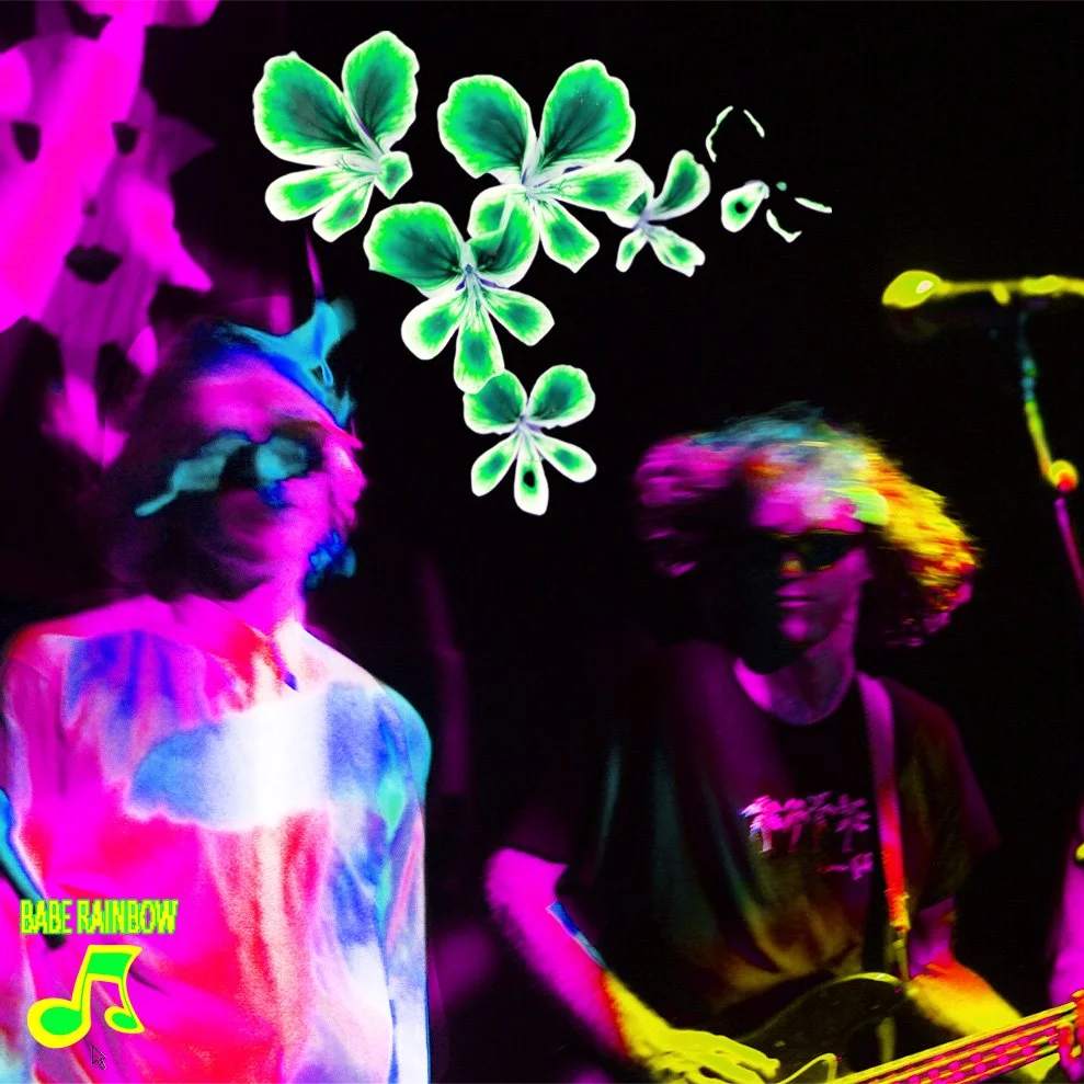 Two musicians performing on stage under blacklights with neon-colored decorations, including green four-leaf clovers and a pink rainbow logo.