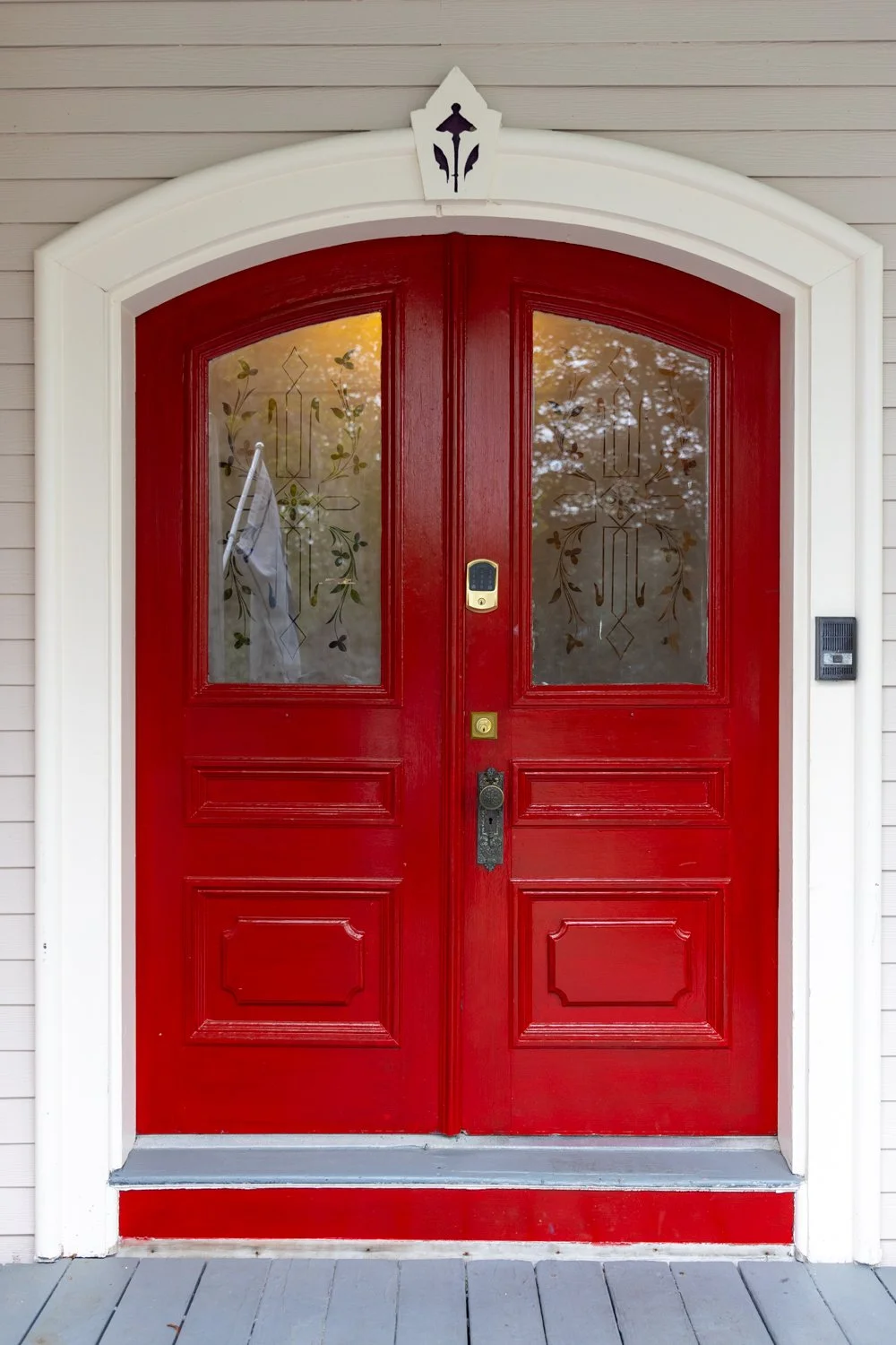 Red front door with glass windows, decorative glass designs, and a white arched frame, mounted on a house with beige siding.