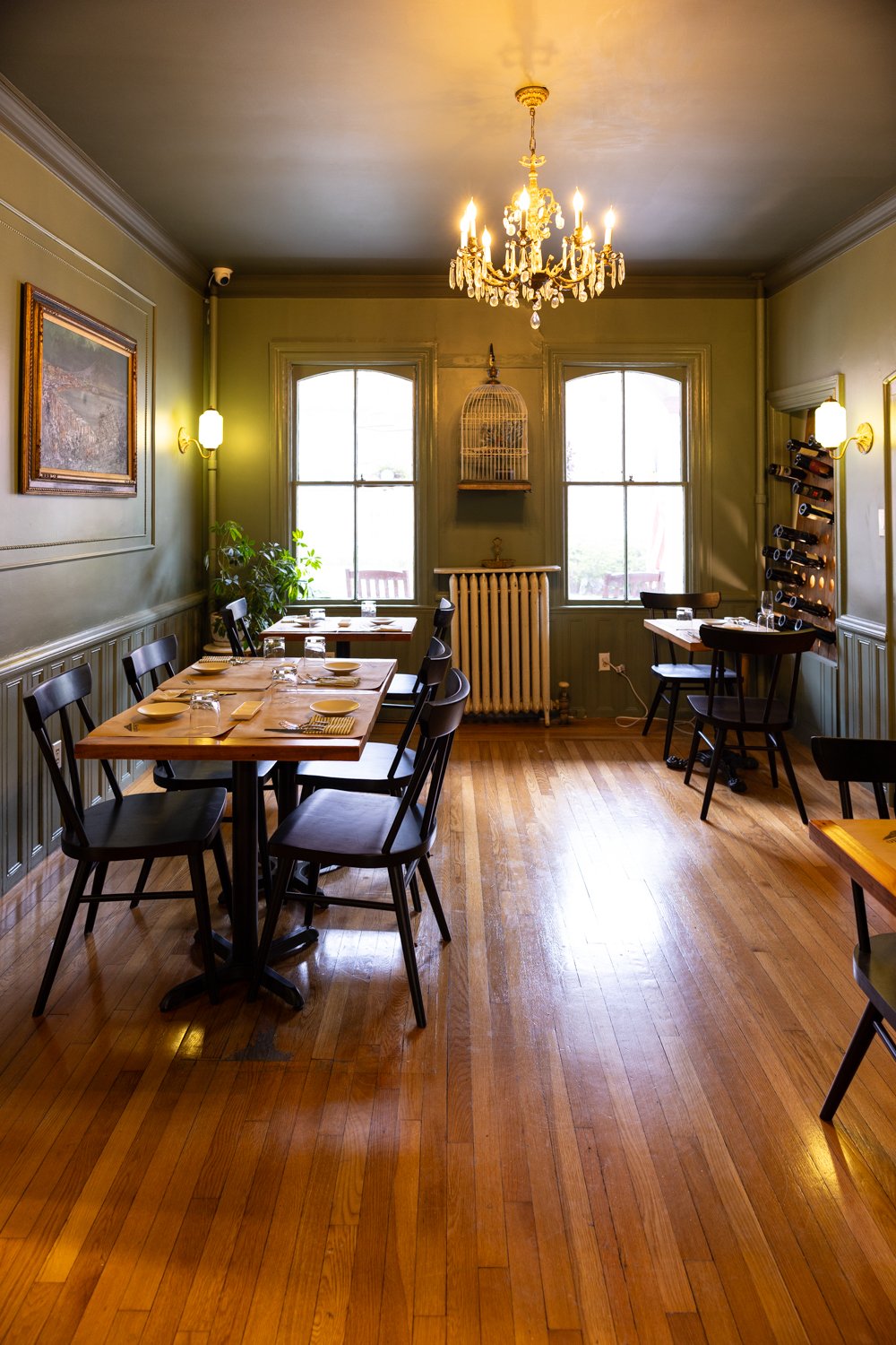 A cozy restaurant dining room with a chandelier, large windows, wooden flooring, and tables set with plates and glasses.
